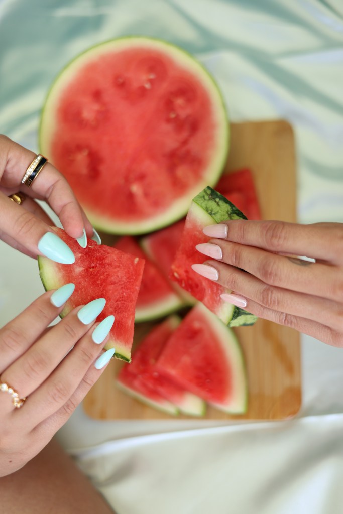 Two hands reaching for slices of watermelon on a wooden cutting board, with one hand holding a piece and the other poised to grab another.