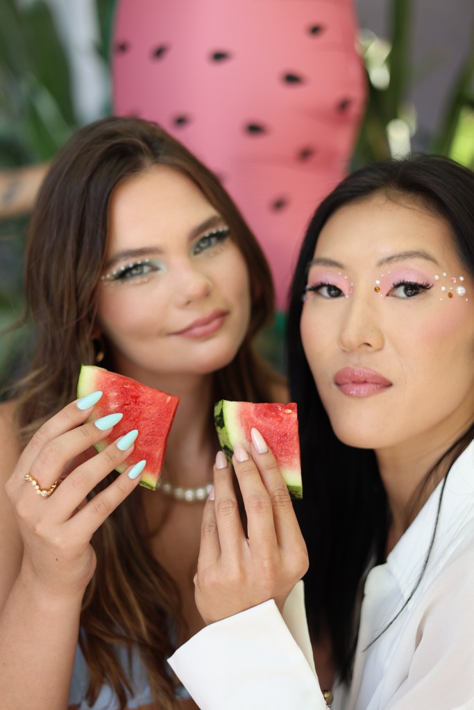 Two women holding slices of watermelon, with vibrant makeup and colorful nails, in a lively, tropical setting.