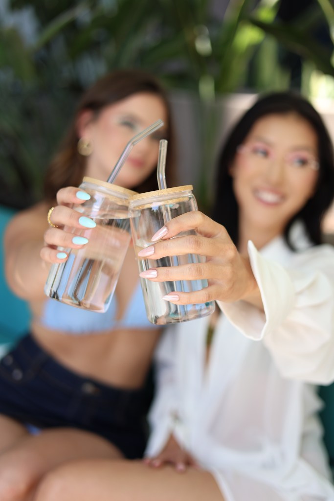Two women holding glass jars filled with water, toasting with bamboo straws, surrounded by greenery.