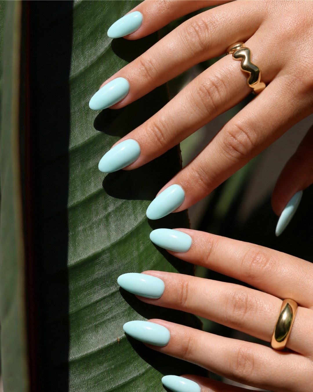Remove the film from the image while preserving the hand, glossy pale sky-blue almond-shaped press-on nails, gold rings, tropical green leaf, composition, lighting, and overall visual style. Do not alter any other elements.